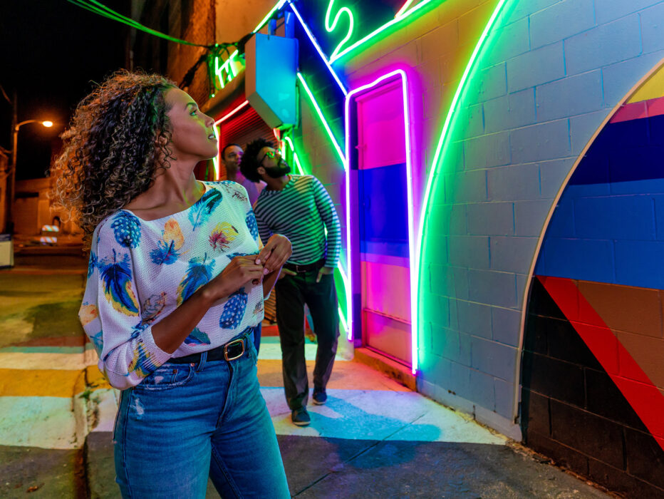 Un grupo de amigos explora de noche el Mural de la Calle Eléctrica, un callejón iluminado por vibrantes luces de neón dispuestas en patrones geométricos sobre una colorida pared pintada.