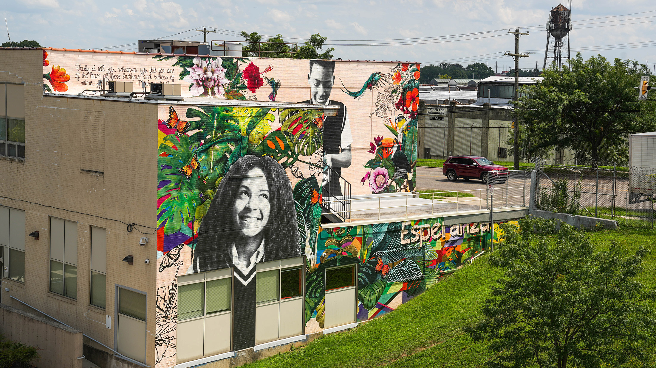 A vibrant mural on the side of a building in Philadelphia features large black-and-white portraits of two young individuals surrounded by lush, colorful tropical plants, flowers, butterflies, and birds.