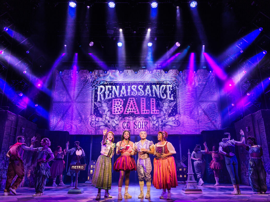 Performers on stage in colorful, Renaissance-inspired costumes sing and dance under vibrant purple and blue stage lighting.