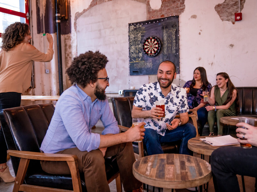 People sit on leather chairs and sofas while enjoying beers at Love City Brewing. A person in the background gets ready to throw a dart at a dartboard on the wall.
