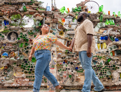 A couple holds hands while walking by a wall covered in artwork featuring reclaimed items like glass bottles and bicycle spokes.