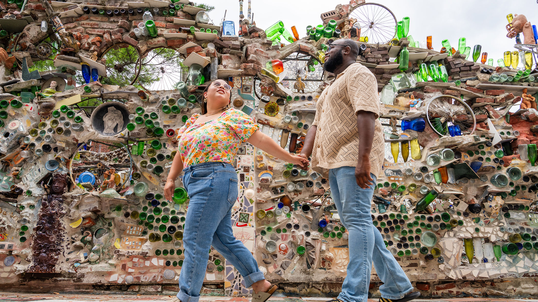 A couple holds hands while walking by a wall covered in artwork featuring reclaimed items like glass bottles and bicycle spokes.