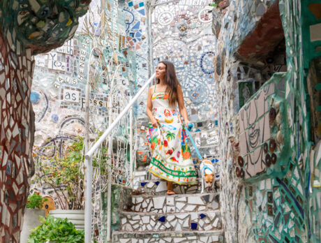 A woman wearing a white dress with a colorful print walks down the mosaic stairs at Philadelphia's Magic Gardens. She holds a teal leash attached to a small brown and white dog.