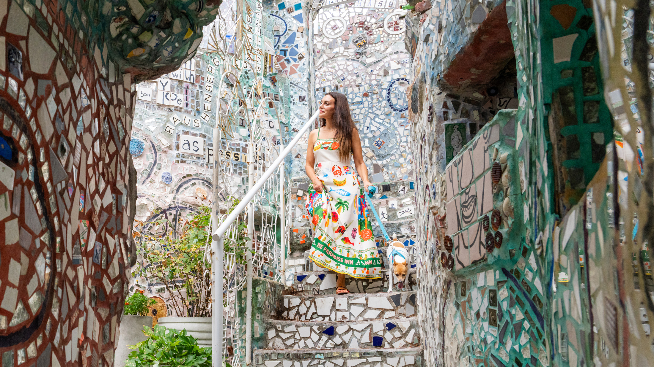 A woman wearing a white dress with a colorful print walks down the mosaic stairs at Philadelphia's Magic Gardens. She holds a teal leash attached to a small brown and white dog.
