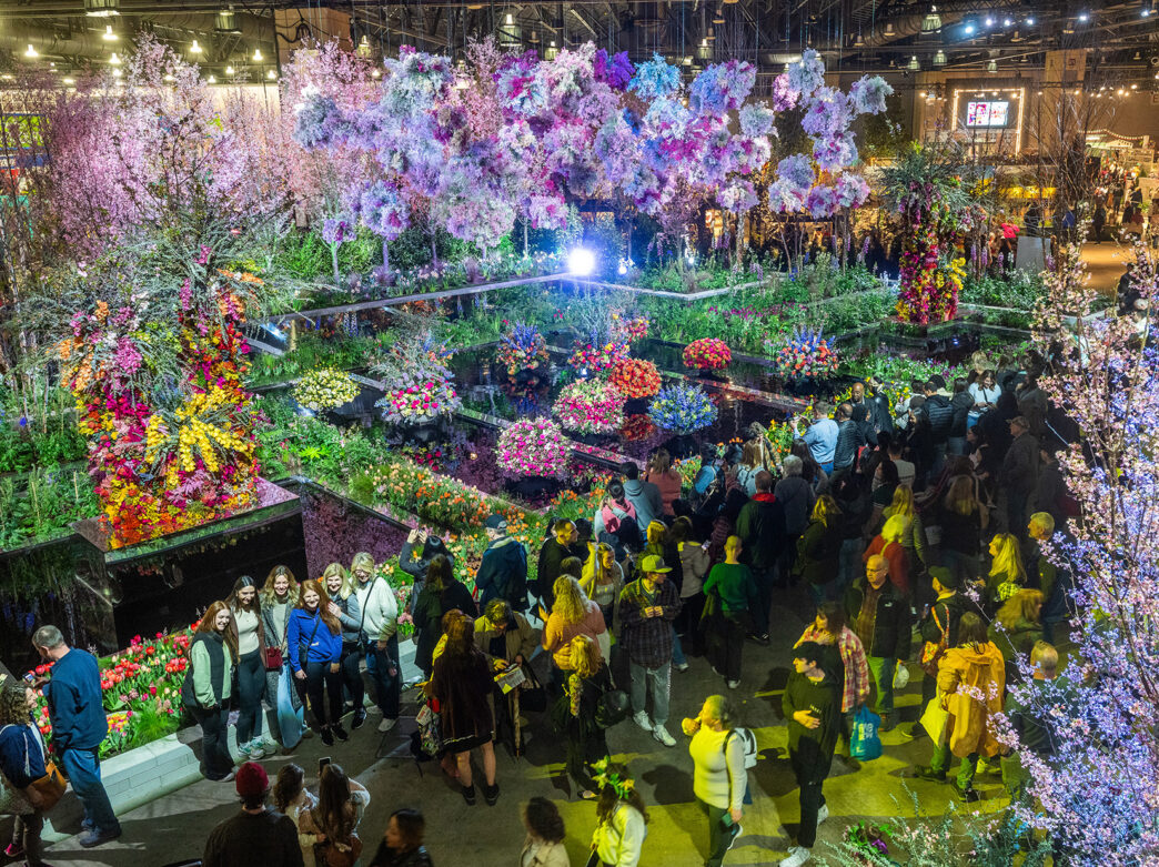 Crowds of people walk through the exhibit hall at the Philadelphia Flower Show. Colorful flower displays hang overhead and float on water