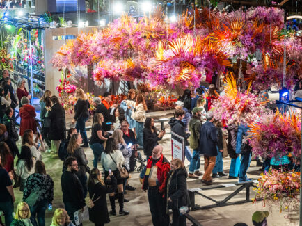 People walk through the exhibit hall at the Philadelphia Flower Show. Pink and orange flower arrangements hang from the ceiling.