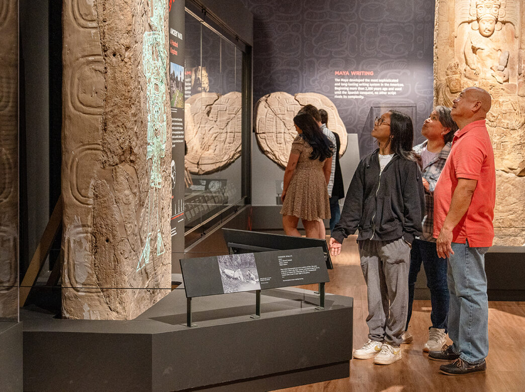 Three museum visitors explore the Mexico and Central America Gallery at the Penn Museum. Various artifacts and sculptures are displayed, including an Mayan alter.