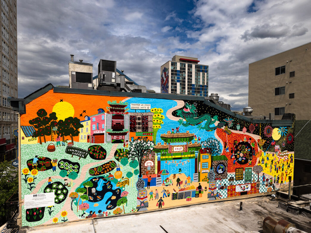 A vibrant mural in Philadelphia's Chinatown showcases a colorful depiction of community life, blending urban and cultural elements with traditional Chinese motifs. The artwork features peopole, a community garden, local rowhomes, the famous Friendship Gate, a green dragon and fireworks.