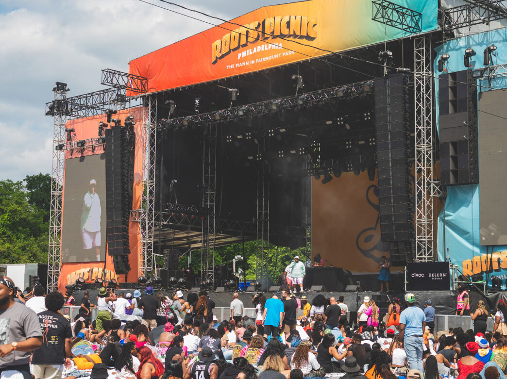 Une foule nombreuse se rassemble devant la scène principale colorée du Roots Picnic, où se produit un artiste vêtu d'une tenue vert clair et coiffé d'une casquette blanche.