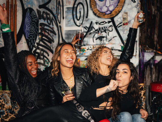 Un grupo de mujeres grita y vitorea frente a la pared de estilo punk de Tattooed Mom.