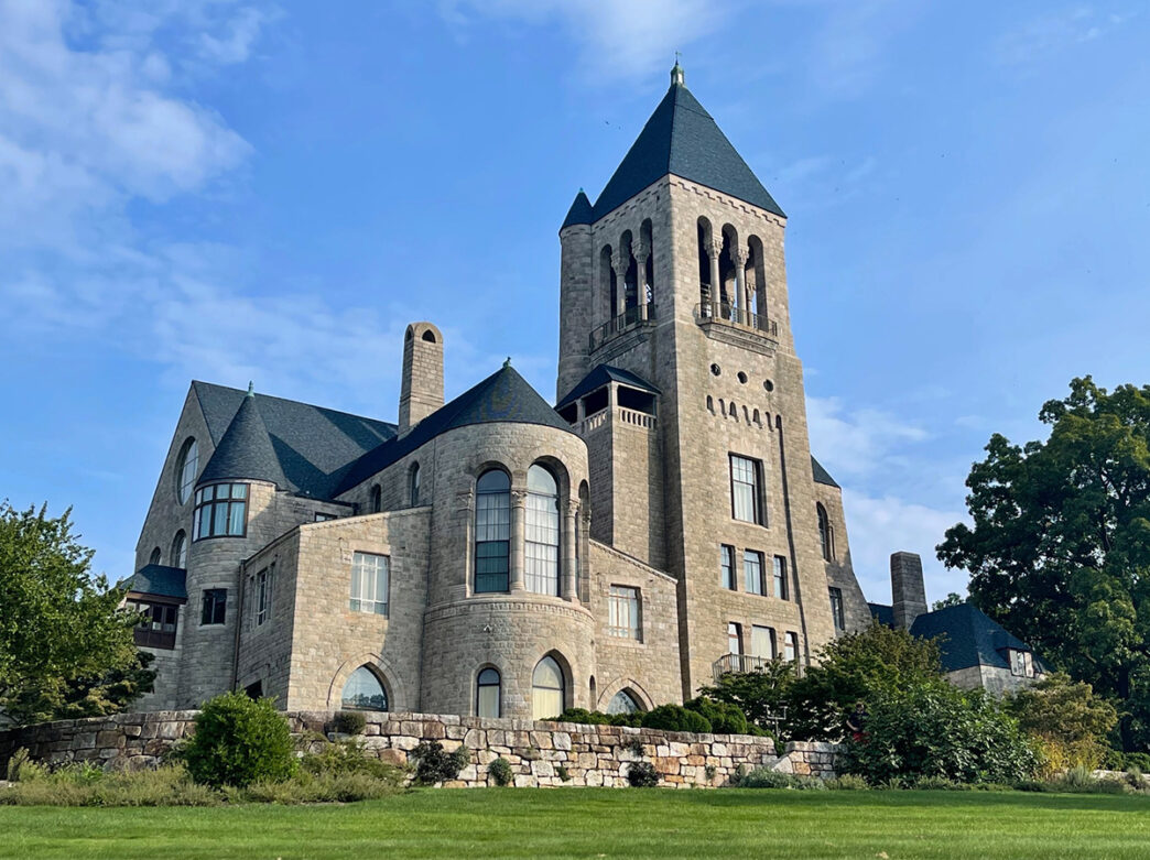 A stone Gilded Age-era building is surrounded by green grass and trees against a blue sky.