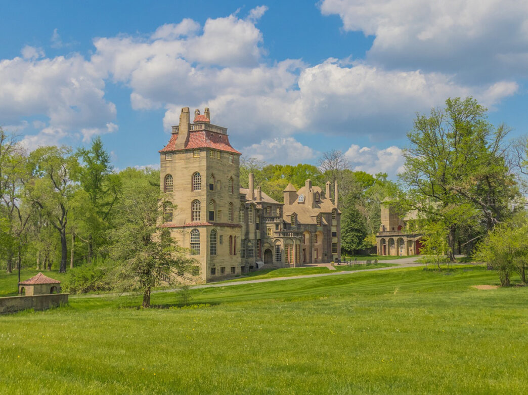 A tan castle with a red roof sits in a field of green grass and trees. The sky is blue with white fluffy clouds above.