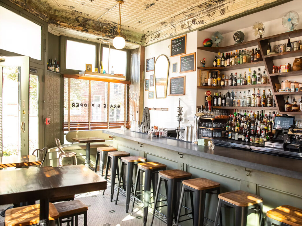 The bar space at Grace and Proper. A long bar with eight stools faces a wall displaying liquor bottles. A few high-top tables and a booth are on the side of the room.