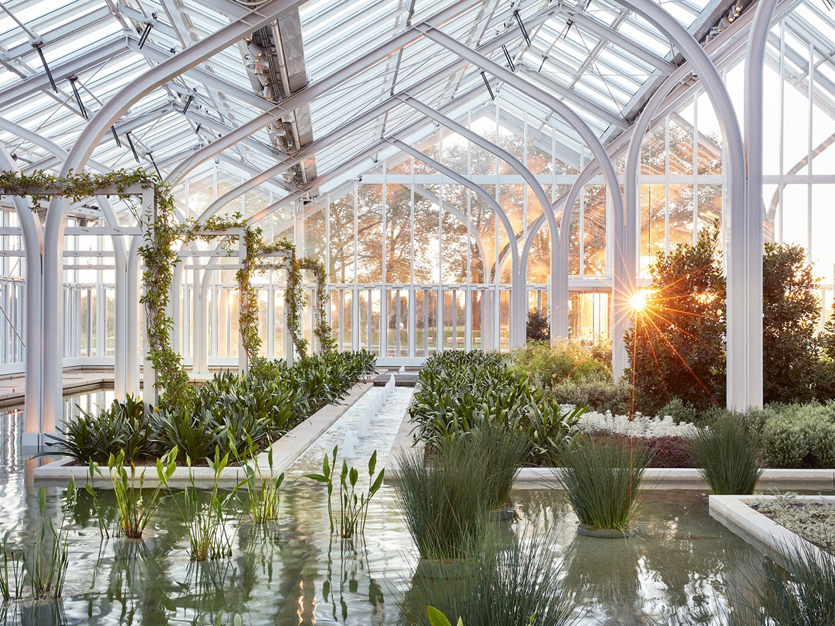 Vista interior do novo Conservatório Oeste em Longwood Gardens com um raio de sol brilhando através do vidro. Várias plantas e fontes de água preenchem a estufa de vidro.
