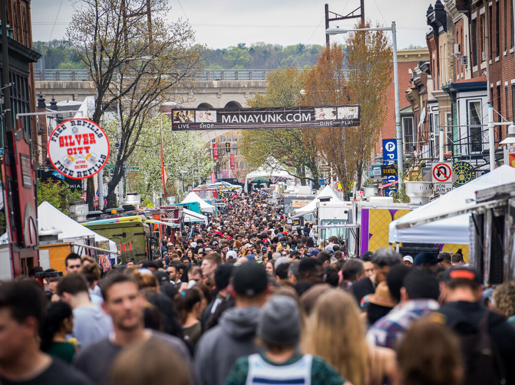 A crowd of people walk through Manayunk during the StrEAT Food Festival. Food trucks and food tents line the street.