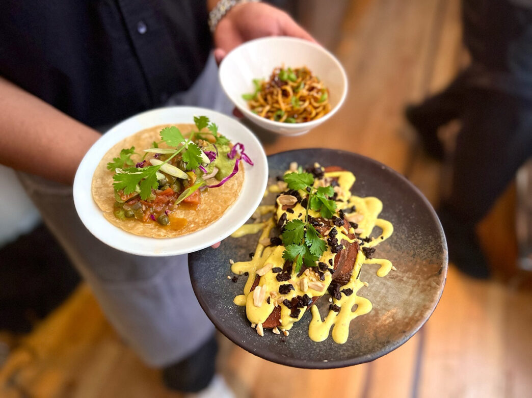Un camarero sostiene tres platos con tacos de coliflor, patatas al ají amarillo y fideos fríos en Vedge.