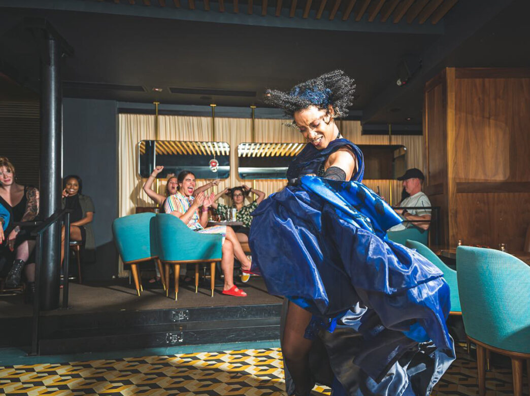A performer wearing a blue dress twirls around in R&D Cocktail Bar while people sitting at the bar and in booths cheer.