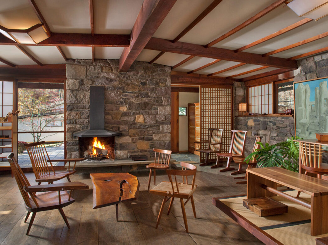The interior of a the showroom at George Nakashima Woodworkers. The room features a wood burning fireplace, various wood chairs and a live edge coffee table. Wood beams run along the ceiling.