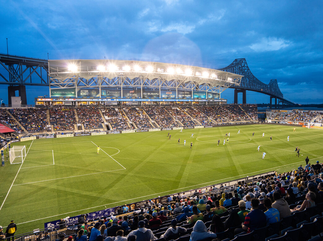 The Philadelphia Union soccer team plays a game in the evening. The sky is dark but Subaru Park is illuminated with stadium lighting. A bridge over the Delaware River is seen in the background.