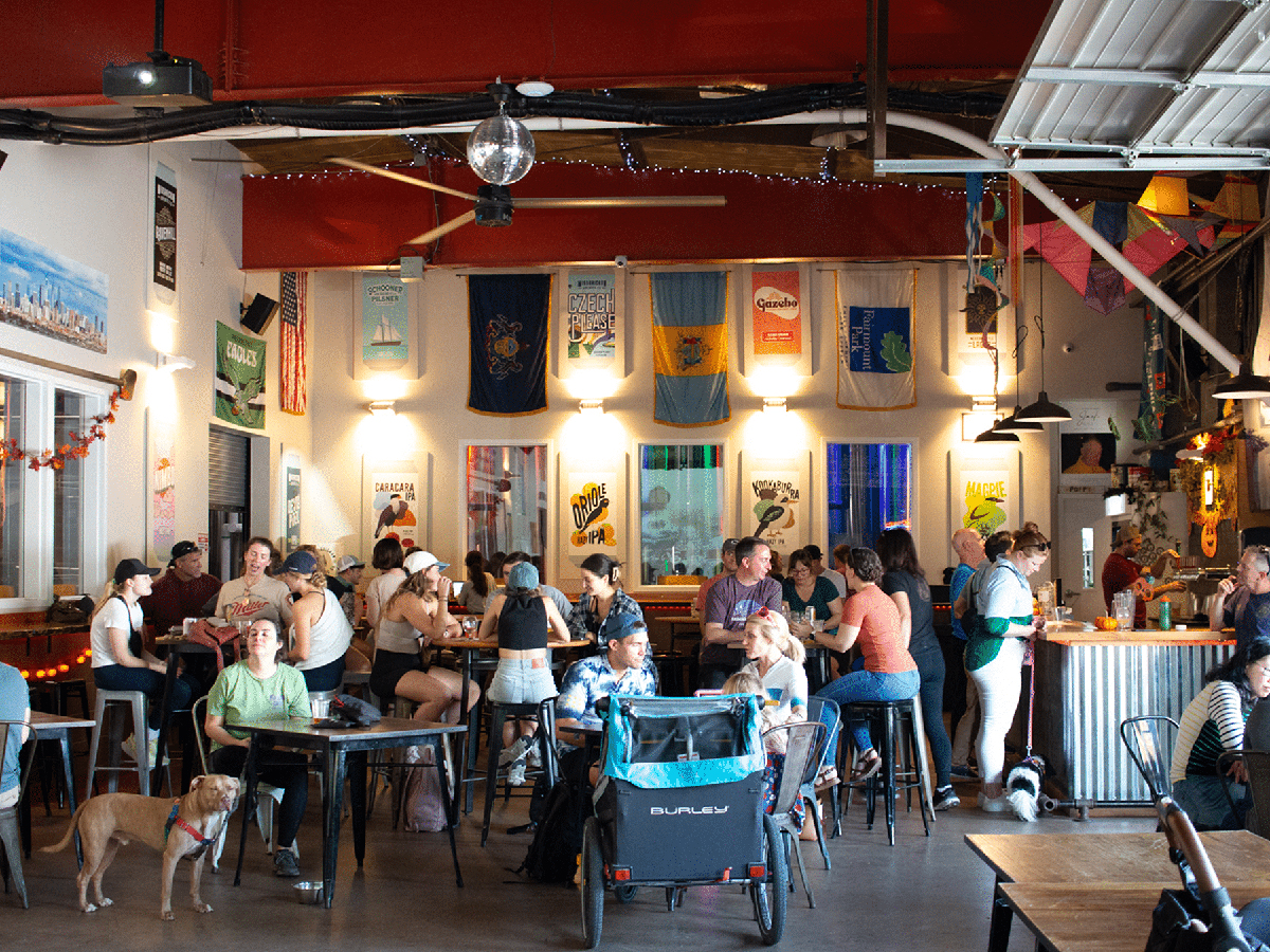 People sit at high top tables and regular tables at Wissahickon Brewing Company's tasting room. Two people have dogs on leashes. Flags hang from the back wall and a disco ball hangs from the ceiling.