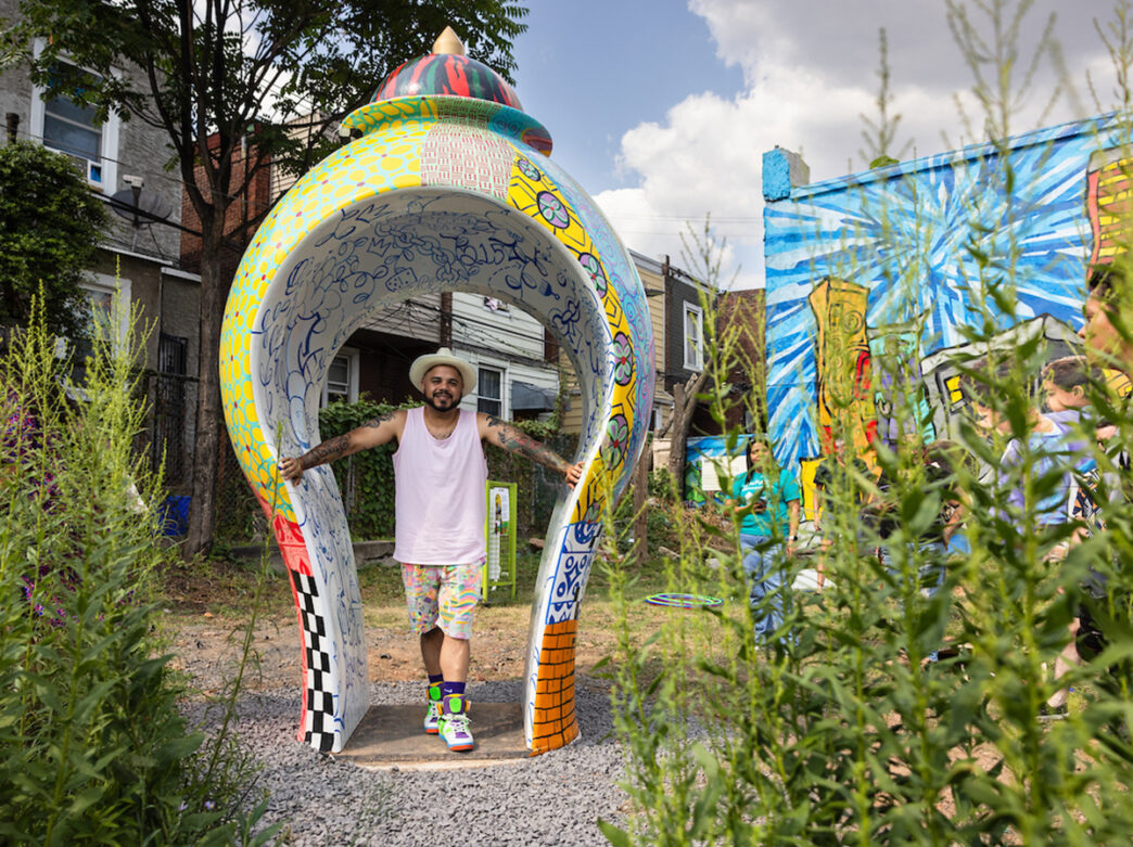Artist Roberto Lugo stands under an arch-shaped vessel at the Kensington Corridor Trust. The piece of art, We Here, features a colorful mosaic of patterns.