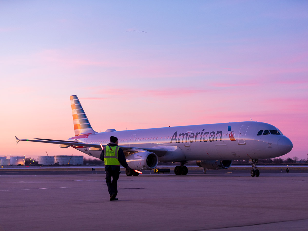 A ground crew member in a reflective vest walks toward an American Airlines aircraft that has landed on the tarmac at sunrise.