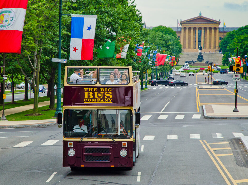 A double-decker open-air tour bus drives down Benjamin Franklin Parkway with the Philadelphia Museum of Art in the background. The parkway is lined with green trees and flags from around the world.