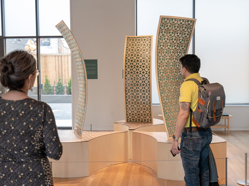 Two people look at a clay installation on display at the Clay Studio in Philadelphia. The installation includes 3 curved panels on elevated platforms.