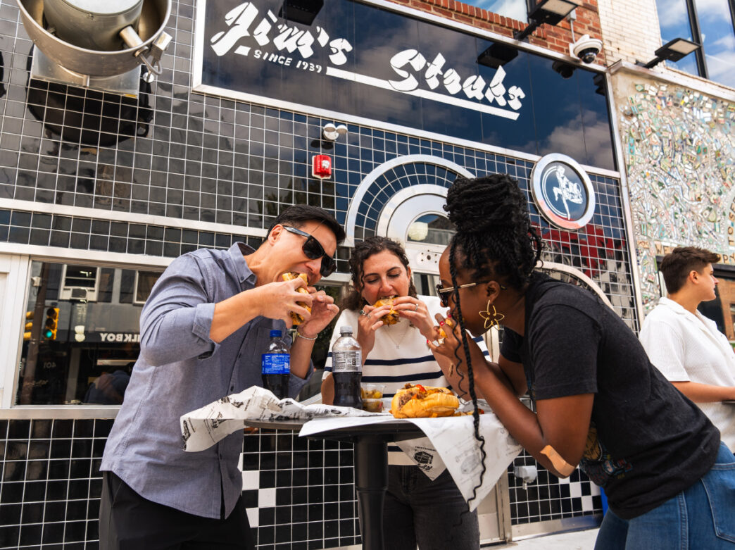 Three people stand at a high-top table outside of Jim's Steaks. Each person bites into a cheesesteak.