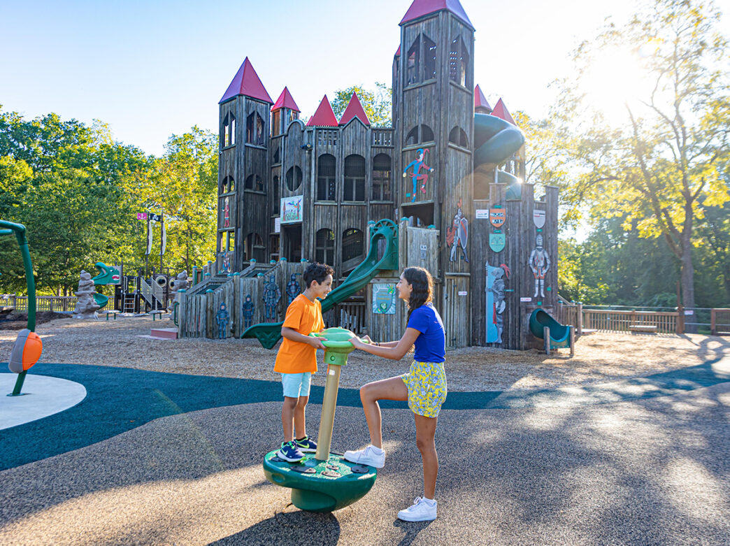 Two children play on an interactive structure in front of Kids Castle, a medieval-themed wooden playground featuring slides, towers and climbing areas in a park setting.