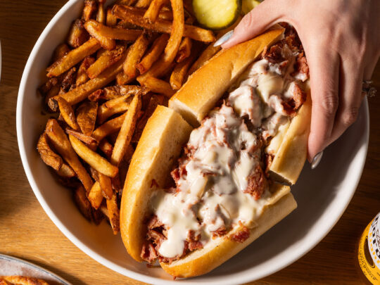 A hand with silver painted nails reaches for half a a cheesesteak on a round plate with crispy French fries and pickle slices. The Wit or Witowski Kielbasa Cheesesteak includes chipped kielbasa with onions and melted American cheese on a long roll.