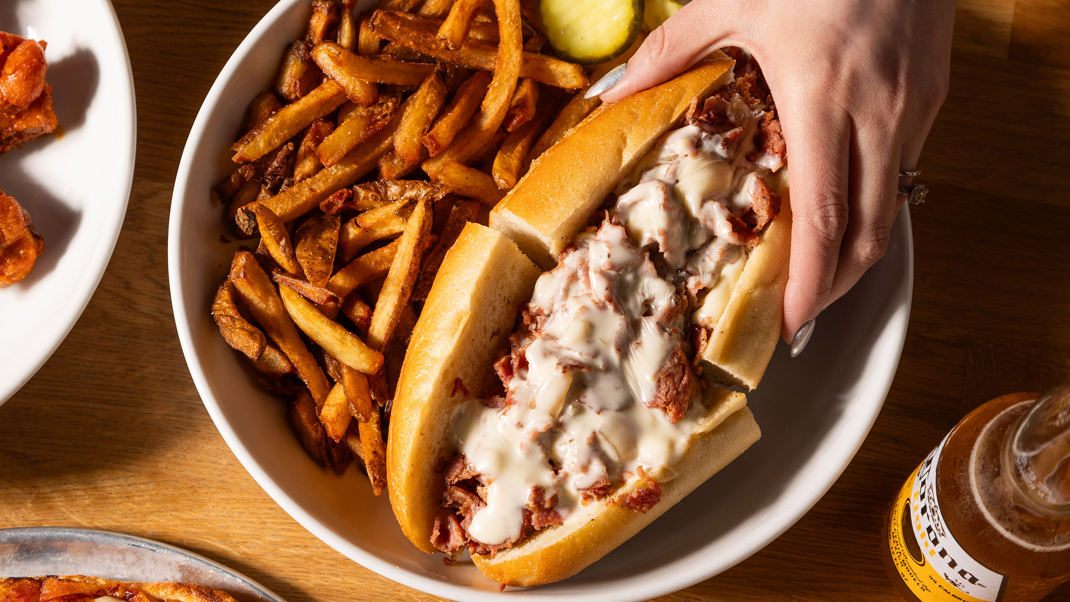 A hand with silver painted nails reaches for half a a cheesesteak on a round plate with crispy French fries and pickle slices. The Wit or Witowski Kielbasa Cheesesteak includes chipped kielbasa with onions and melted American cheese on a long roll.