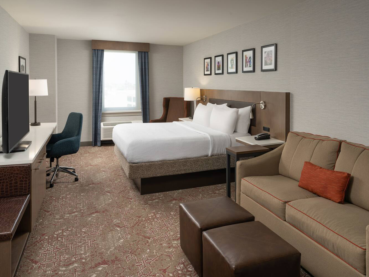 A king bed, couch with footrests and desk fill a guest room at Hilton Garden Inn Philadelphia Center City. The bed is made with white linens and the room has brown carpeting.