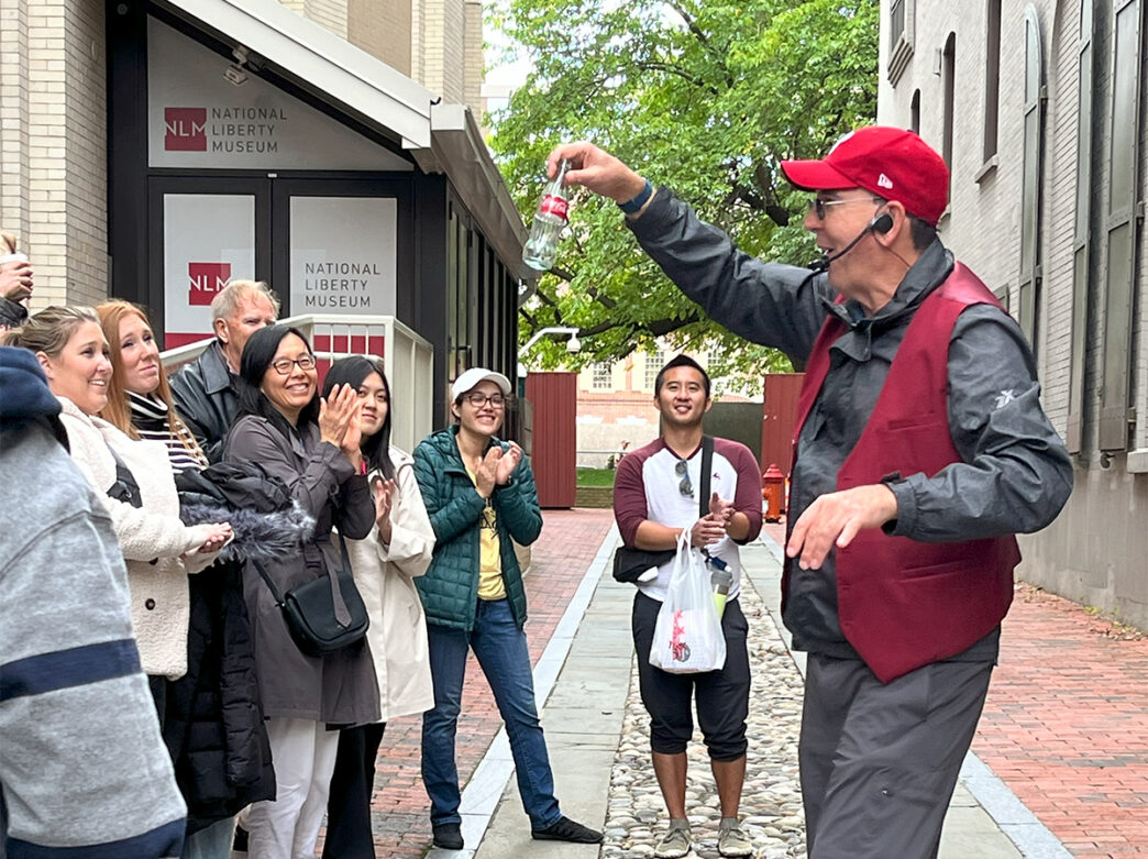 A man wearing a red baseball hat and red vest holds up a glass Coca-Cola bottle during a magic trick while the tour group around him claps.