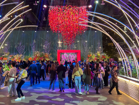 A crowd of people walk between foliage and arched neon lights with red cascading flowers overhead at the PHS Philadelphia Flower Show