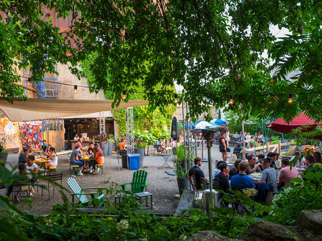 People socialize and relax at an outdoor gravel patio with umbrellas and awnings. Green trees and plants surround the outdoor space.