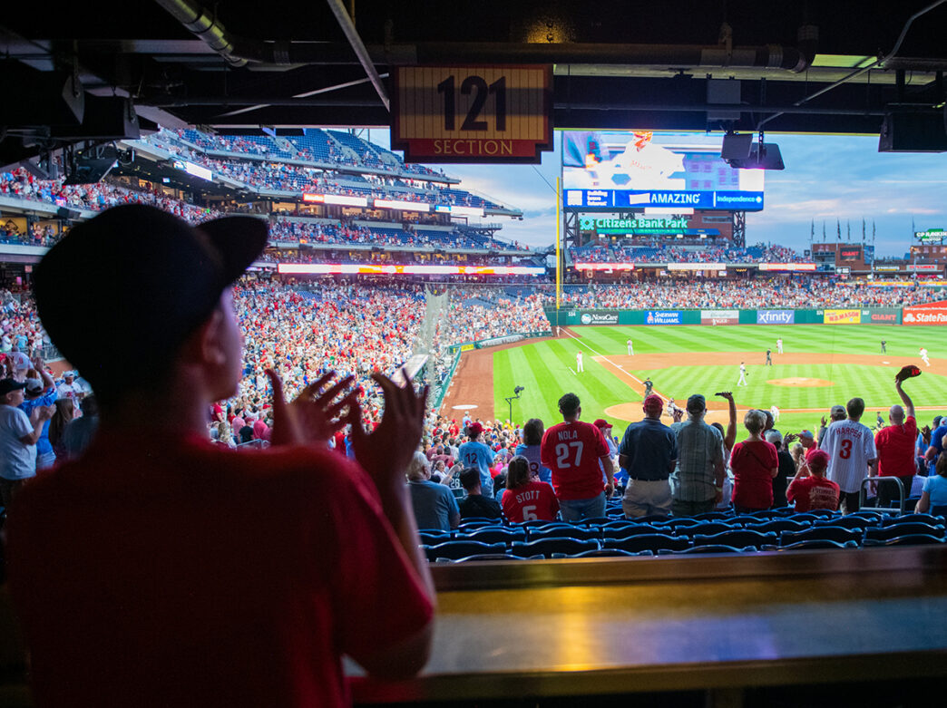 Un jeune fan qui regarde le match depuis une tribune ombragée du Citizens Bank Park applaudit avec enthousiasme, se joignant à la foule animée qui encourage les Phillies.