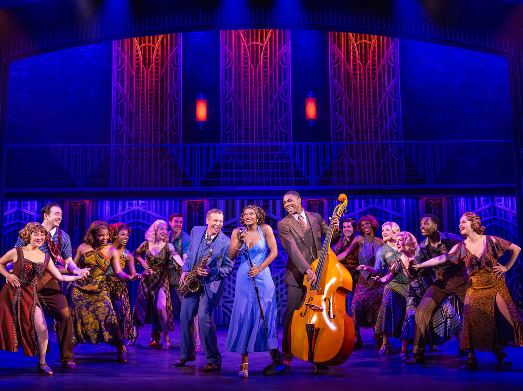A woman wearing a blue dress holds onto a vintage microphone and two men wearing suits lean in towards her. One man has a saxophone and one plays a upright bass. Other cast members surround them during a production of Some Like It Hot.