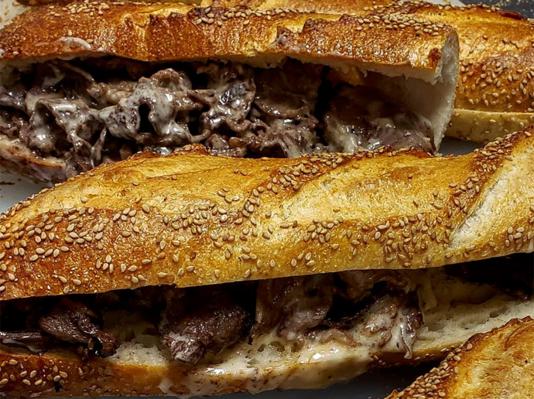 A detail image of a cheesesteak on a seeded roll. Melted cheese drips over sliced beef.