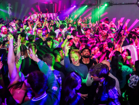 People dance on a crowded dance floor illuminated with green, pink and purple lighting. People raise their hands in the air.