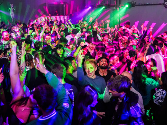 Des personnes dansent sur une piste de danse bondée, éclairée par des lumières vertes, roses et violettes. Les gens lèvent les mains en l'air.