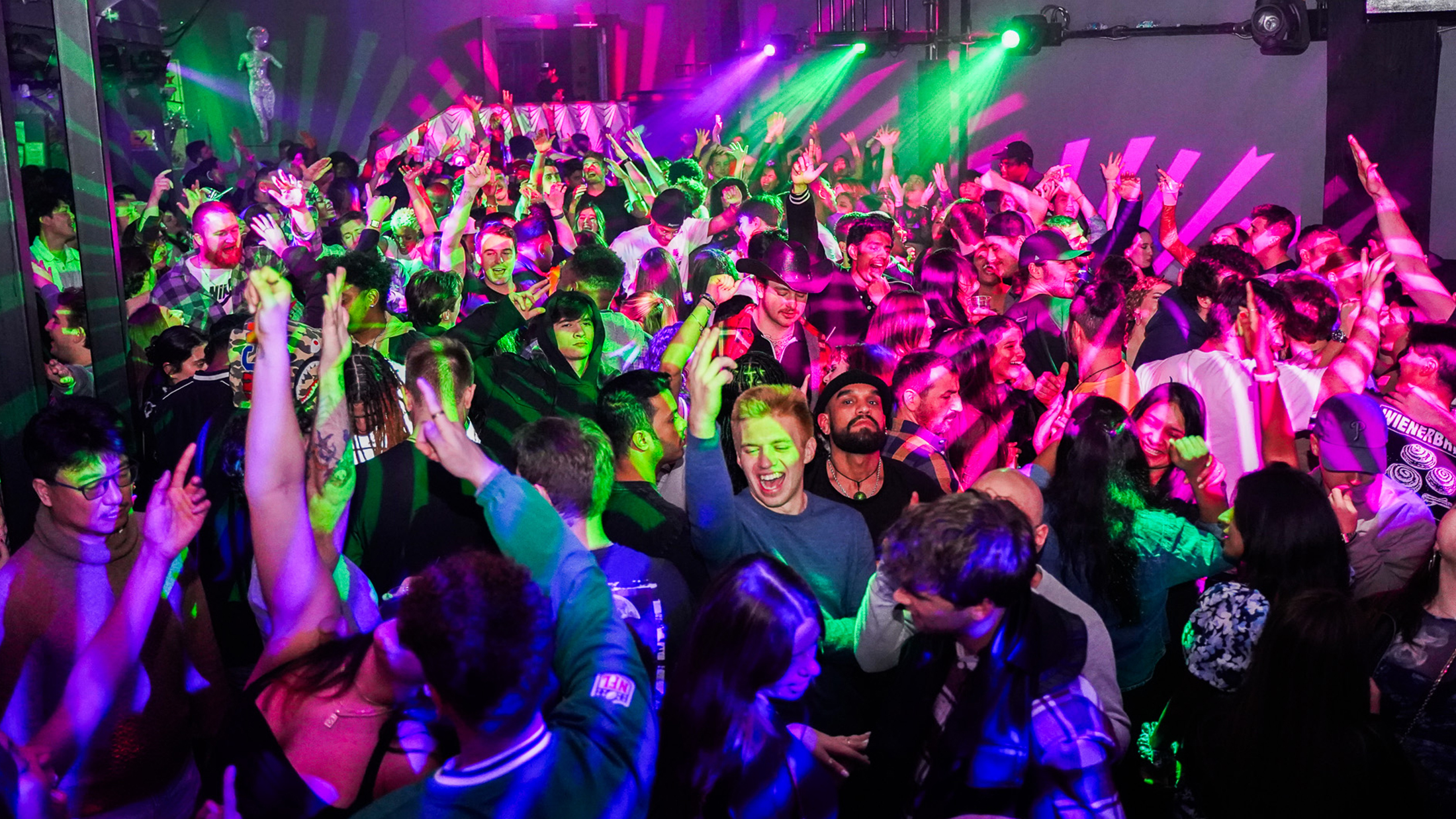 People dance on a crowded dance floor illuminated with green, pink and purple lighting. People raise their hands in the air.