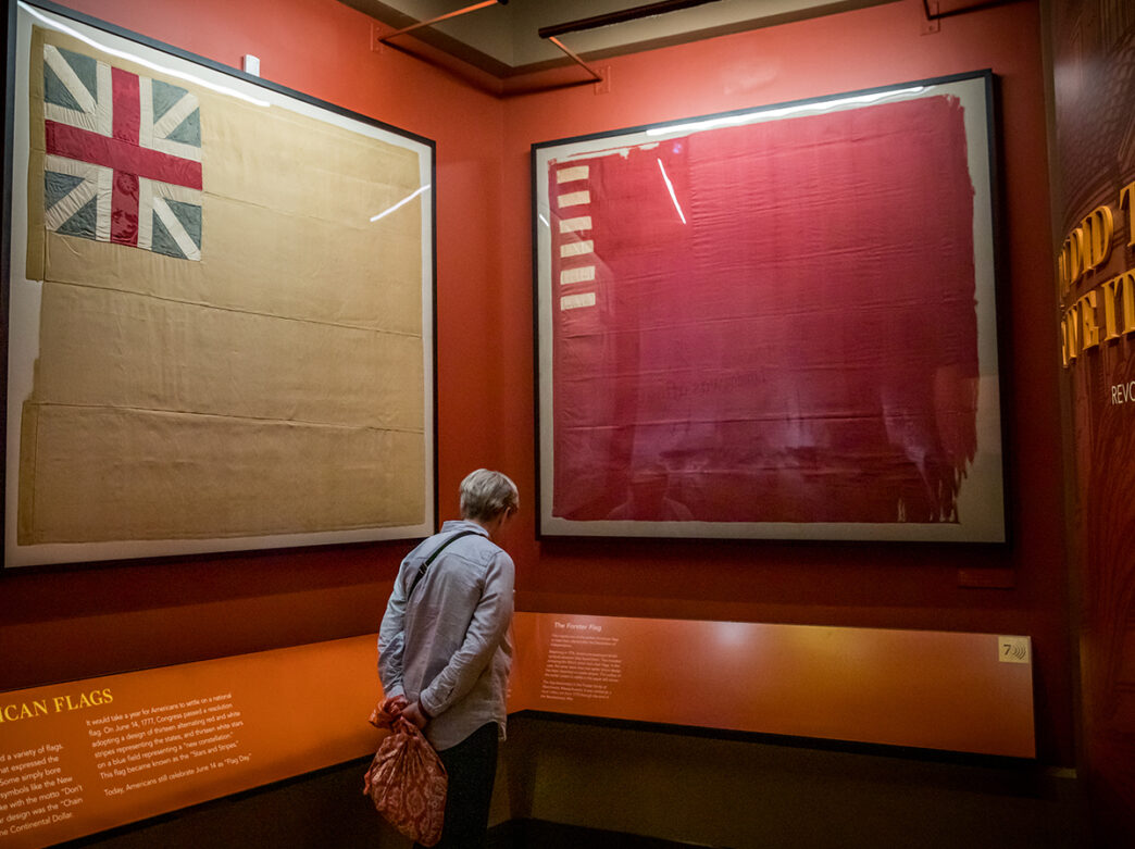 Un visitante examina grandes banderas históricas enmarcadas expuestas en una pared roja del Museum of the American Revolution.