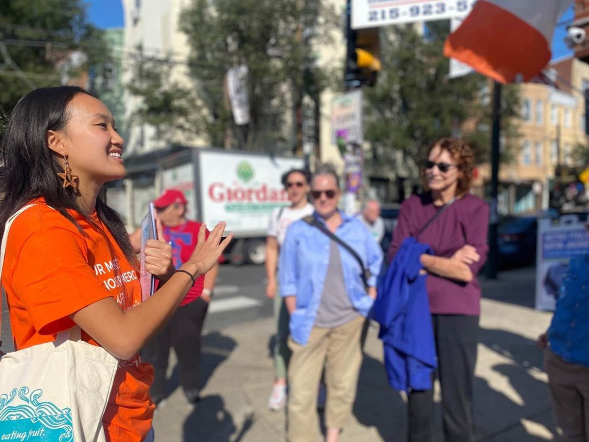 A tour guide wearing an orange t-shirt talks to a group while leading an Our Market Tour along South 9th Street in Philadelphia.