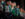 A lively group of dancers in shimmering green and silver costumes performs a high-energy Irish dance routine, smiling as they execute synchronized kicks on stage.