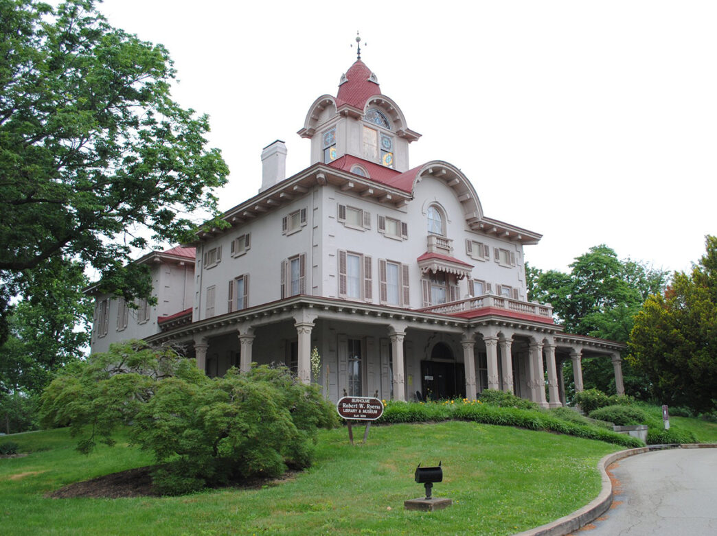 A Victorian mansion with a red roof and wrap around porch set in a green lawn.