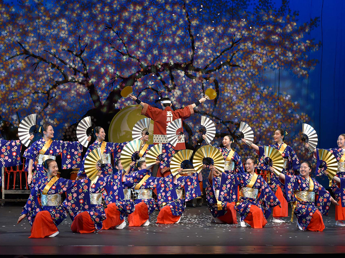 A vibrant performance by Tamagawa University dancers in traditional Japanese attire, holding decorative fans against a backdrop of cherry blossoms.