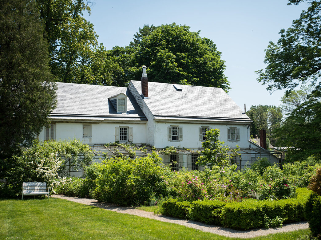 A white historical home with shutters is surrounded by a lush green garden and tall trees at Wyck House and Gardens.