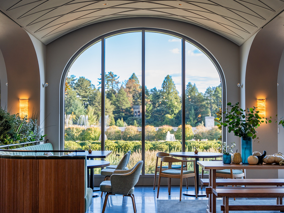 Mesas e cadeiras vazias estão dispostas na sala de jantar de 1906 no Longwood Gardens. As janelas do chão ao teto têm vista para os jardins e fontes.