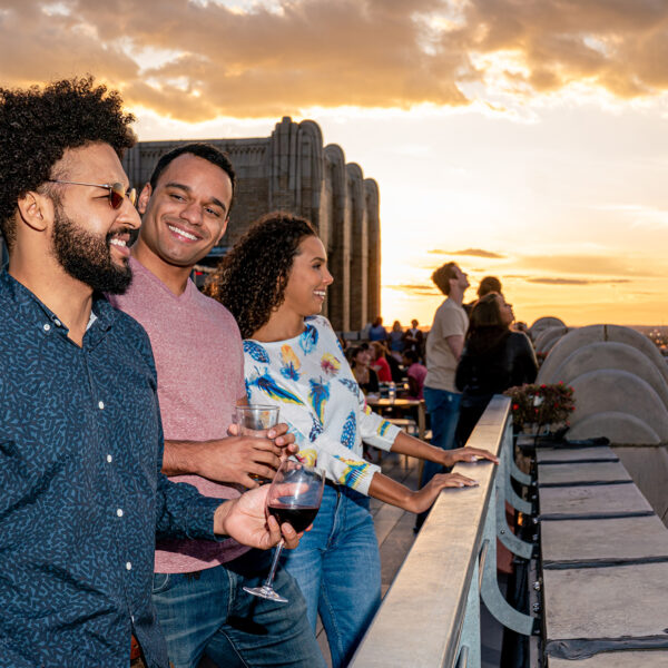 Three people enjoy drinks and look out at the view of the city while at Bok Bar rooftop during sunset. The sky illuminates in yellow and orange tones.